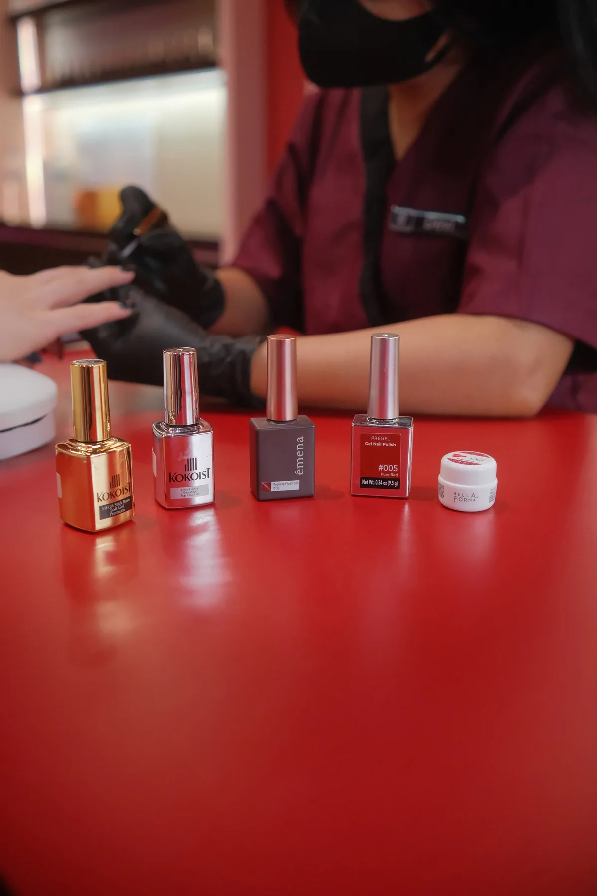 Manicurist applying nail polish to a client. Bottles of nail polish line up on a red table.