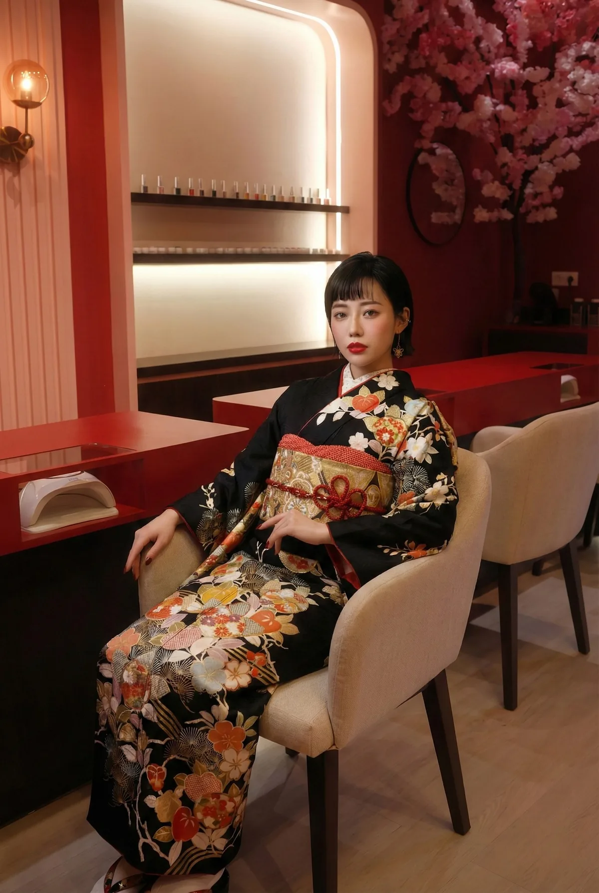 A woman in a beautiful kimono sits in  nail salon, looking at the camera. She's sitting near red surfaces and cherry blossoms.