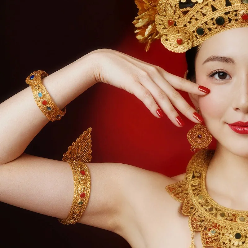 Asian woman in elaborate golden headpiece, jewelry, red lipstick, and manicured nails. Posing hands on cheeks, smiling.